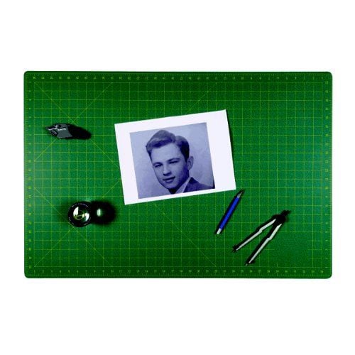 A SpeedPress 23.6" x 35.4" Magic Self-Healing Cutting Mat with a grid pattern, featuring a black-and-white portrait, a utility knife, a magnifying glass, a blue pen, and a pair of compasses neatly arranged on its surface.