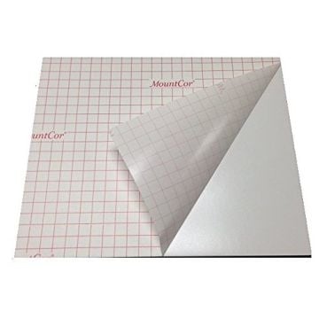A sheet of "MountCor Low Temp Heat Activated Mounting Boards - Black 3/16" Foam" with its adhesive side exposed, featuring a grid pattern and branding, alongside a partially peeled corner revealing the foam backing.